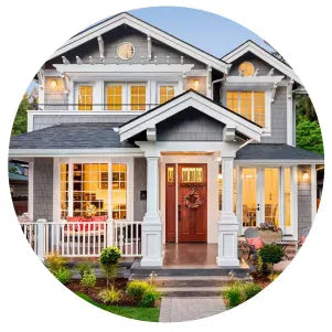 Large modern two-story house with multiple large windows glowing warmly at dusk, white trim, covered front porch, and landscaped yard.