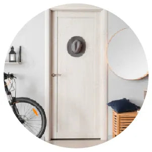 Minimalist white interior door with dark round wreath, modern metal handle, bicycle partially visible on the left, and wooden bench with cushion on the right.