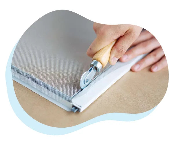 Close-up of a person using a rolling cutter with a wooden handle to trim a mesh screen for a window repair on a flat surface.