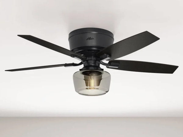 Ceiling-mounted indoor fan with four matte black blades and a clear glass light fixture housing an exposed bulb, set against a white ceiling.