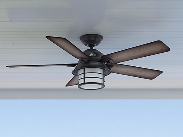 Ceiling-mounted outdoor fan with five weathered wood blades and a round lantern-style light enclosed in a protective cage against a light blue sky.
