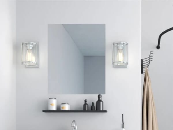 Minimalist bathroom scene featuring a rectangular mirror flanked by two clear glass wall sconces with visible bulbs above a black shelf holding small containers and bottles.
