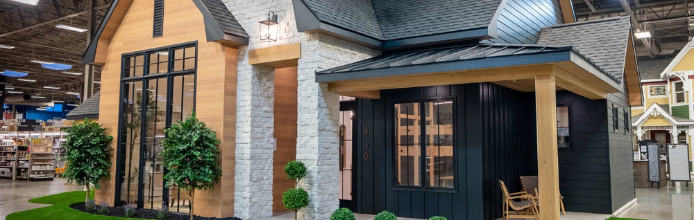 Indoor showroom featuring a detailed small house facade with wood and stone siding, black window frames, and decorative topiary plants