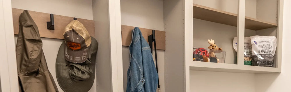 Mudroom storage area featuring light gray built-in cubbies and hooks holding jackets, hats, and bags, with open shelves displaying small decor and supplies.
