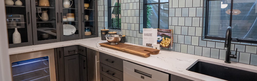 Contemporary kitchen corner with dark lower cabinets, white marble countertop, glass-front upper cabinets displaying dinnerware, and light blue subway tile backsplash under large windows.