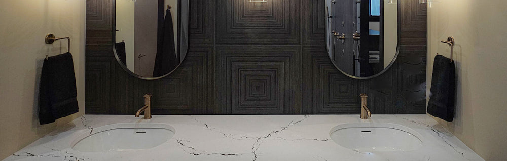 Modern bathroom vanity with dual white marble sinks, sleek gold faucets, round mirrors, dark textured backsplash, and wall-mounted black towels.