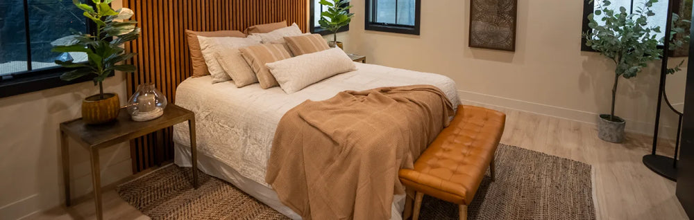 Neutral-toned bedroom with a cream-colored bedspread and tan throw, featuring a leather bench, wood-paneled accent wall, and natural decorative plants.