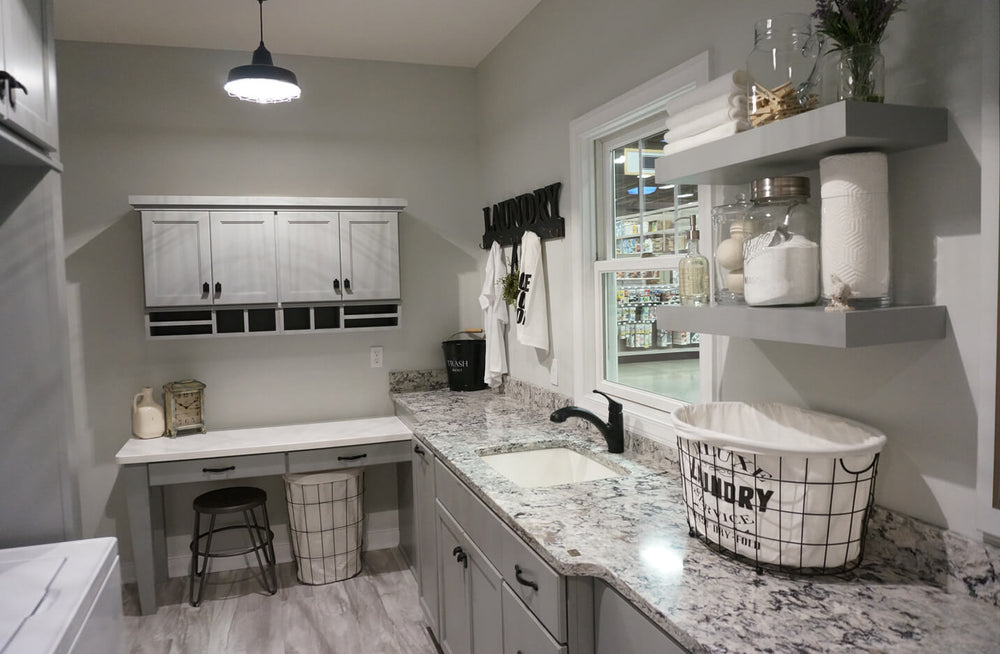 Laundry room featuring light gray cabinets and granite countertops, with an undermount sink, wall shelves holding jars, a stool, and wire baskets for organization.
