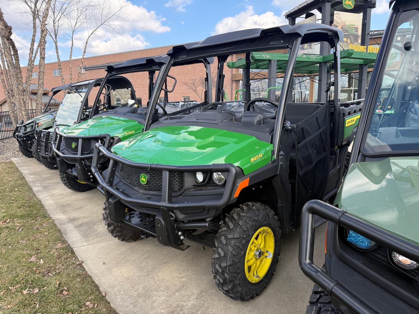 Row of green John Deere crossover utility vehicles with yellow-rimmed tires, protective grills, and roofs displayed outdoors at dealership near other equipment