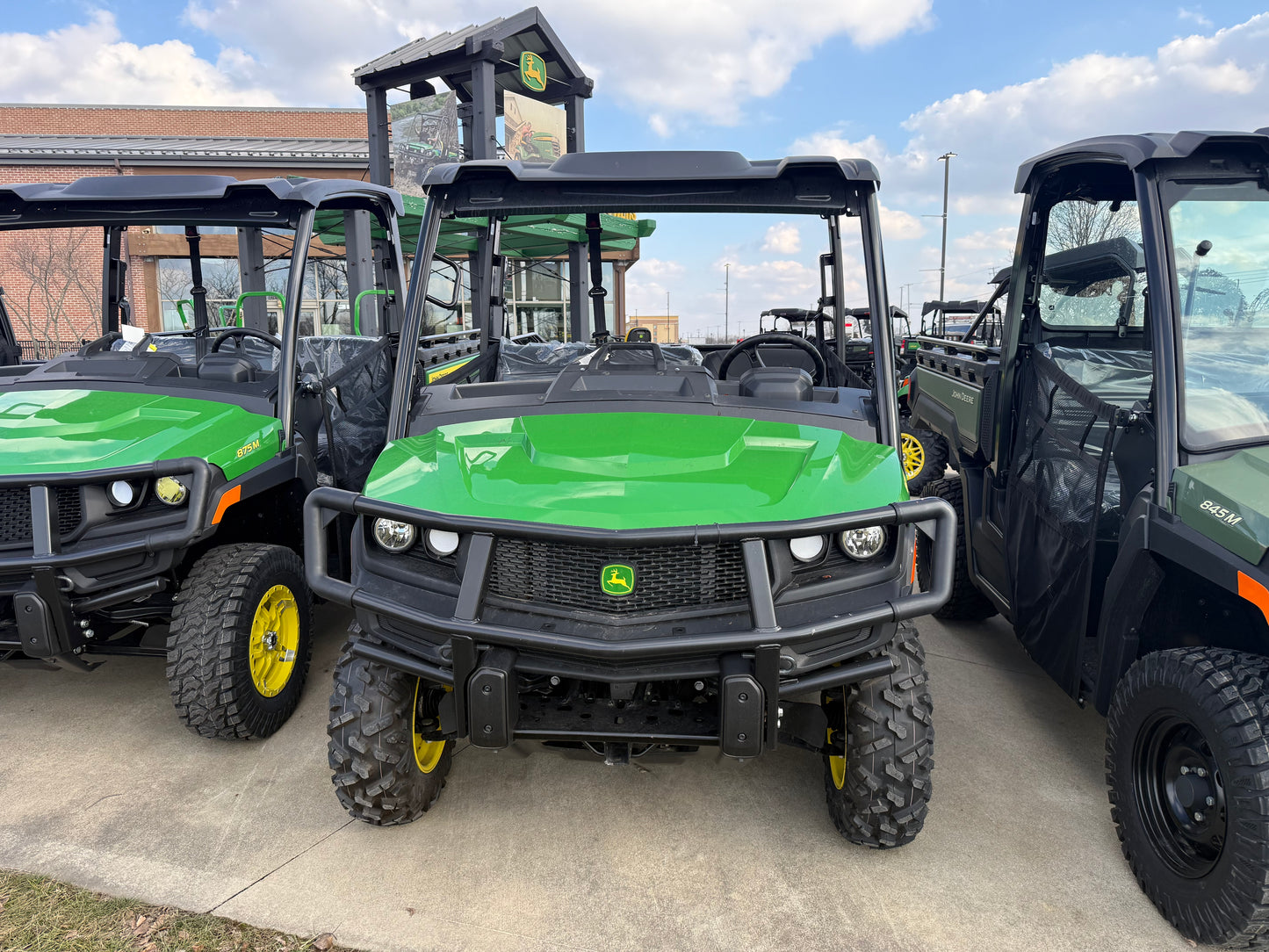 Front view of a green John Deere Custom XUV 845M crossover utility vehicle with rugged off-road tires and protective roof parked outdoors among other John Deere UTVs at dealership lot