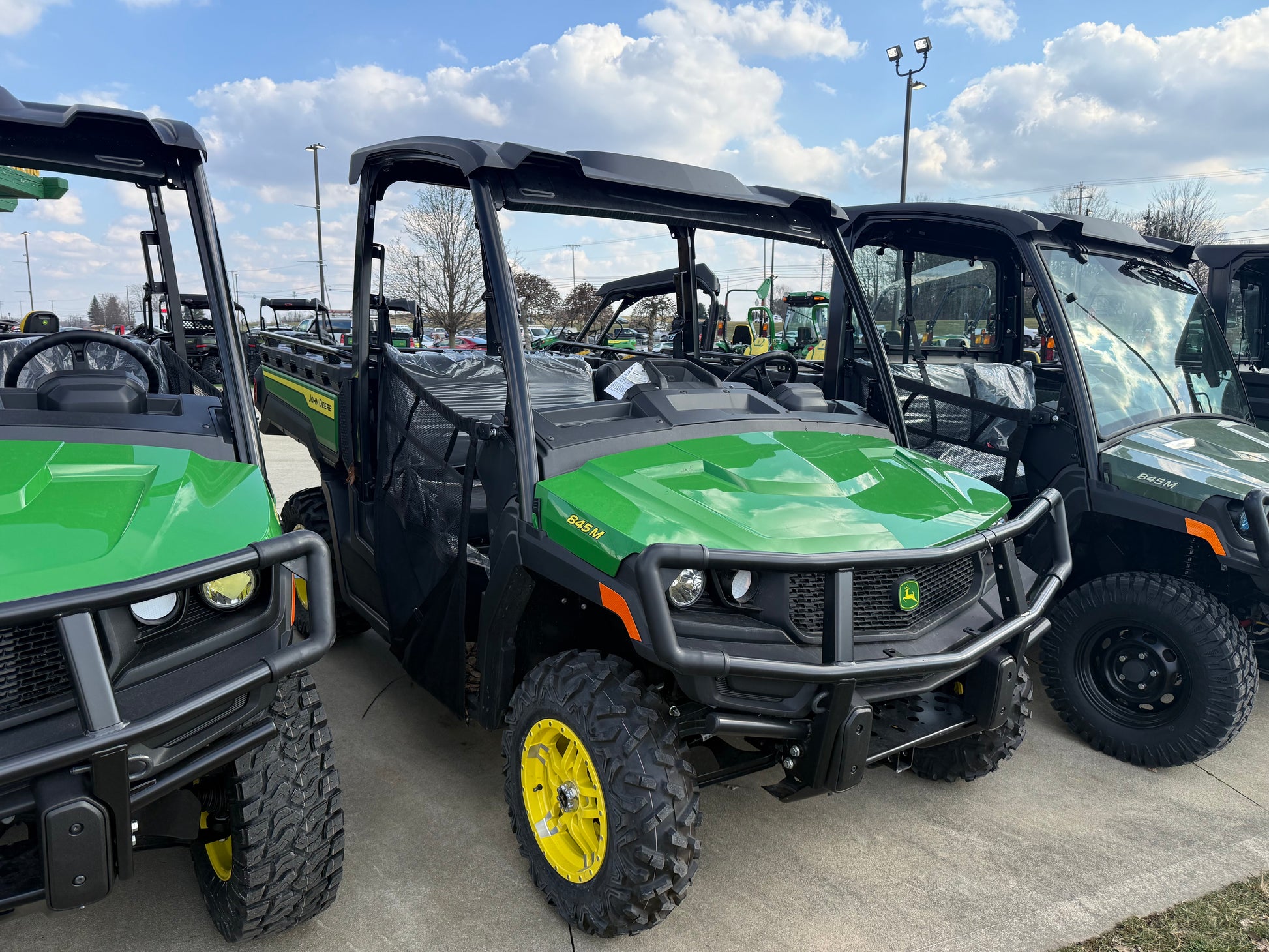 Green John Deere utility vehicle with yellow wheels, protective front grille, and cargo bed parked outdoors on concrete among other equipment at dealership