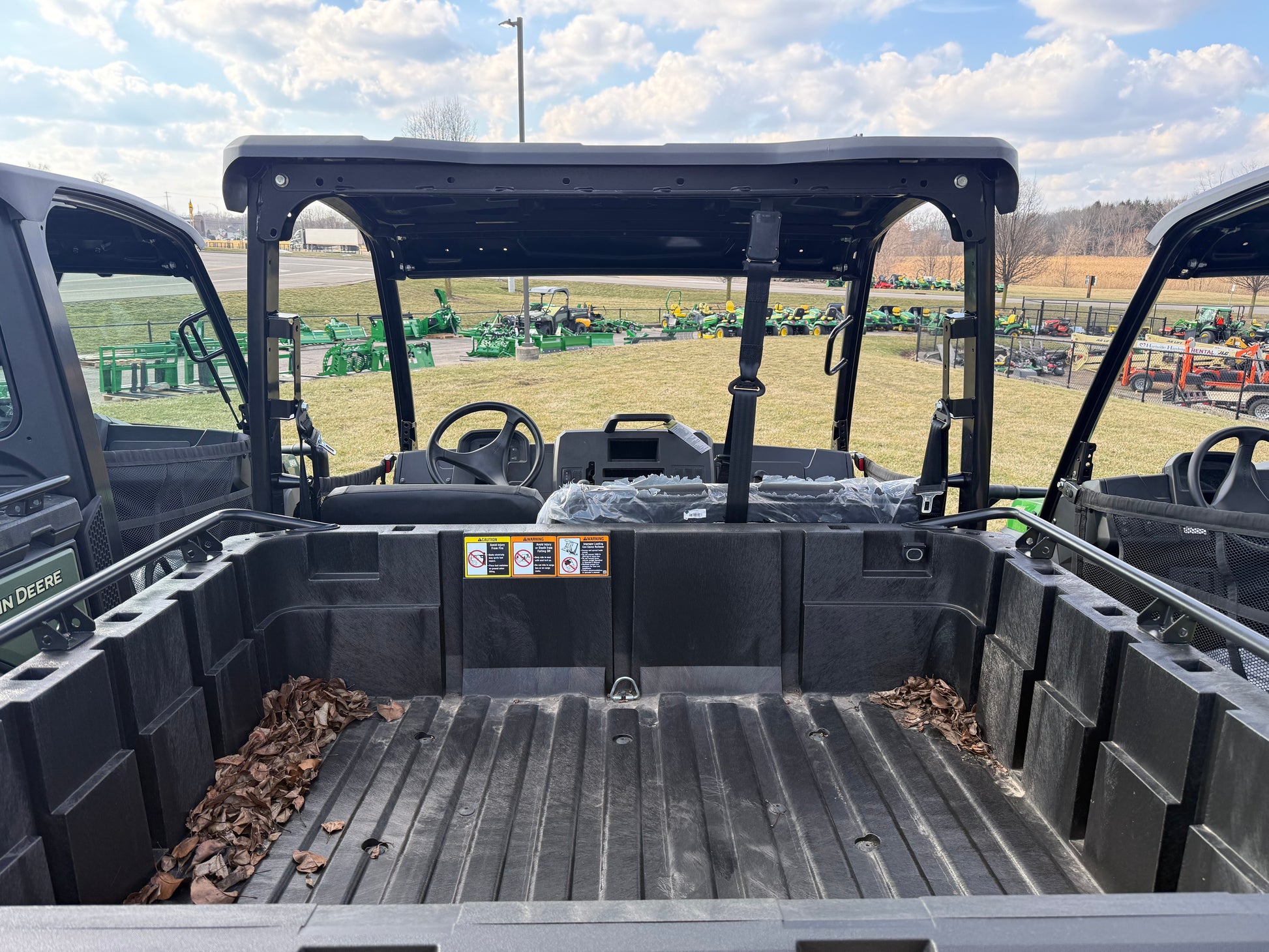 Cargo bed view of a John Deere JD621318 Custom XUV 845M crossover utility vehicle with protective roof and partial interior seen at outdoor dealership lot with agricultural equipment