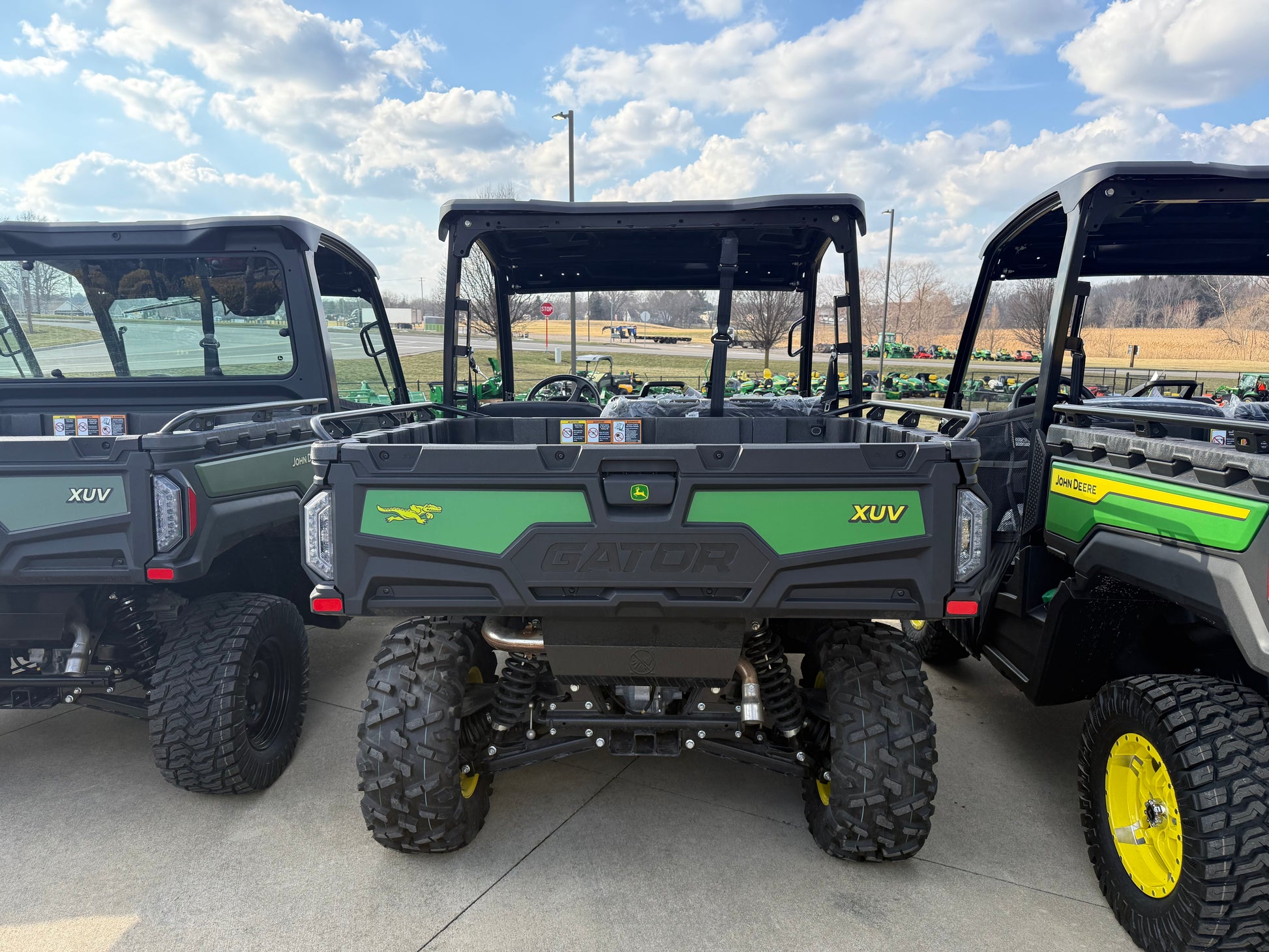 Rear view of three John Deere crossover utility vehicles with rugged tires and cargo beds parked outdoors on a concrete lot among other equipment