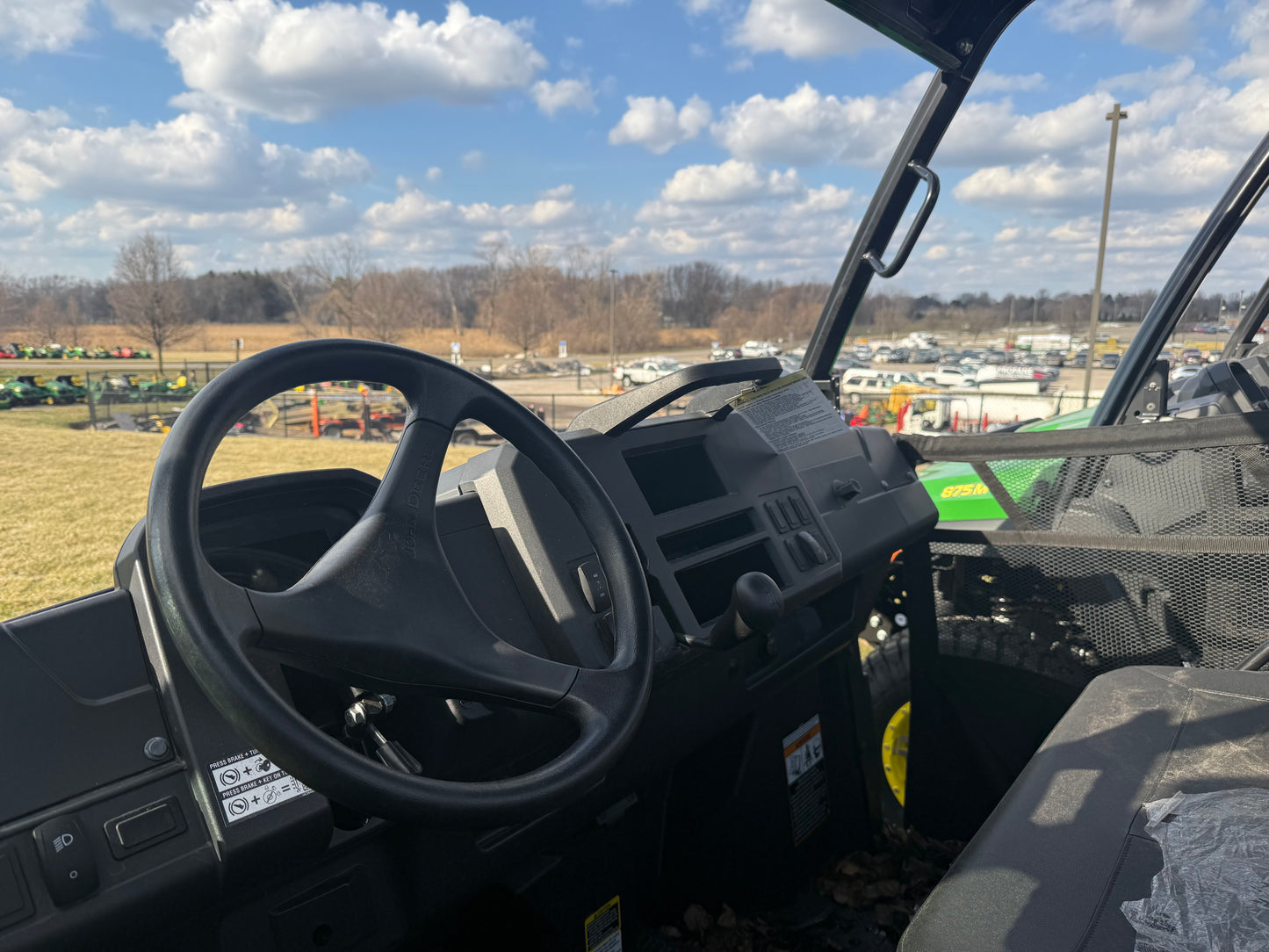 Interior view of a John Deere utility vehicle showing steering wheel, dashboard controls, and bench seating with mesh side door at outdoor equipment lot.