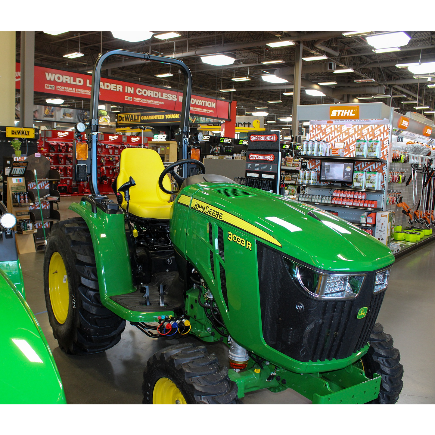 Green John Deere 3033R compact utility tractor with yellow seat and large tread tires displayed indoors in an outdoor power equipment and lawn & garden tools store.