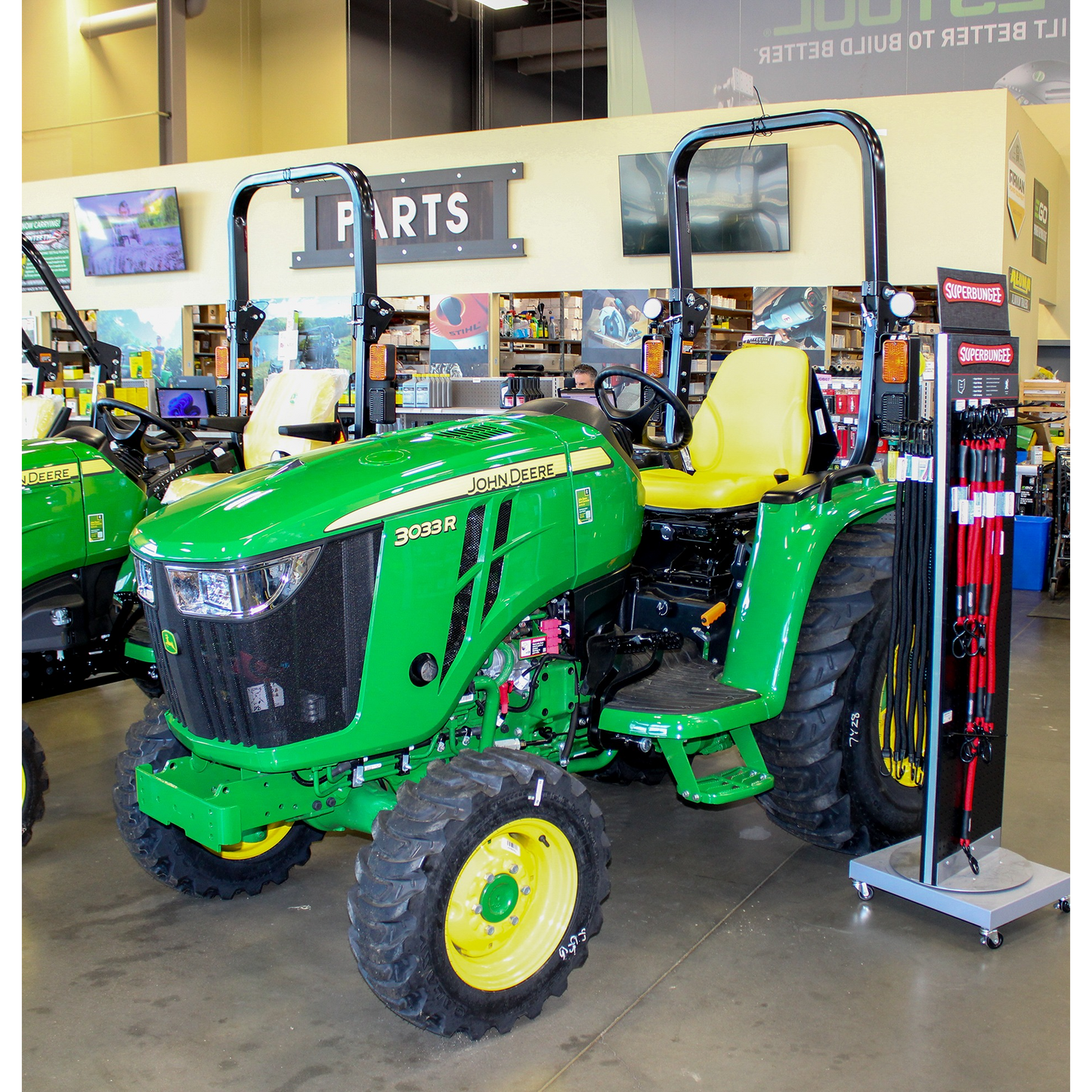 Green John Deere 3033R compact utility tractor with yellow seat and large tread tires displayed indoors among outdoor power equipment and John Deere parts accessories.