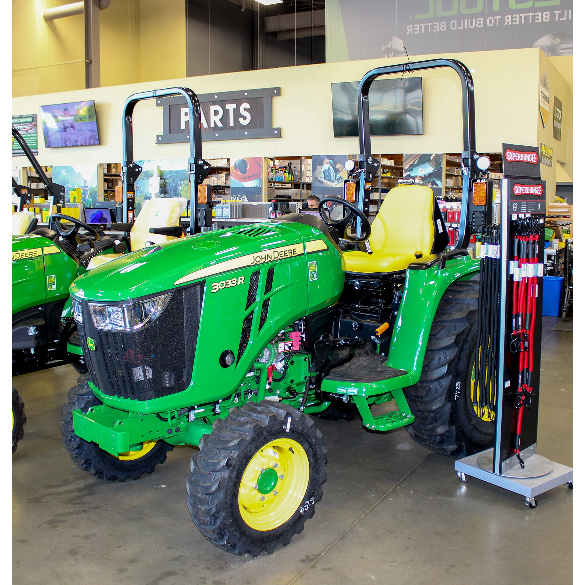 Green John Deere 3033R compact utility tractor with yellow seat and large tread tires displayed indoors among outdoor power equipment and John Deere parts accessories.