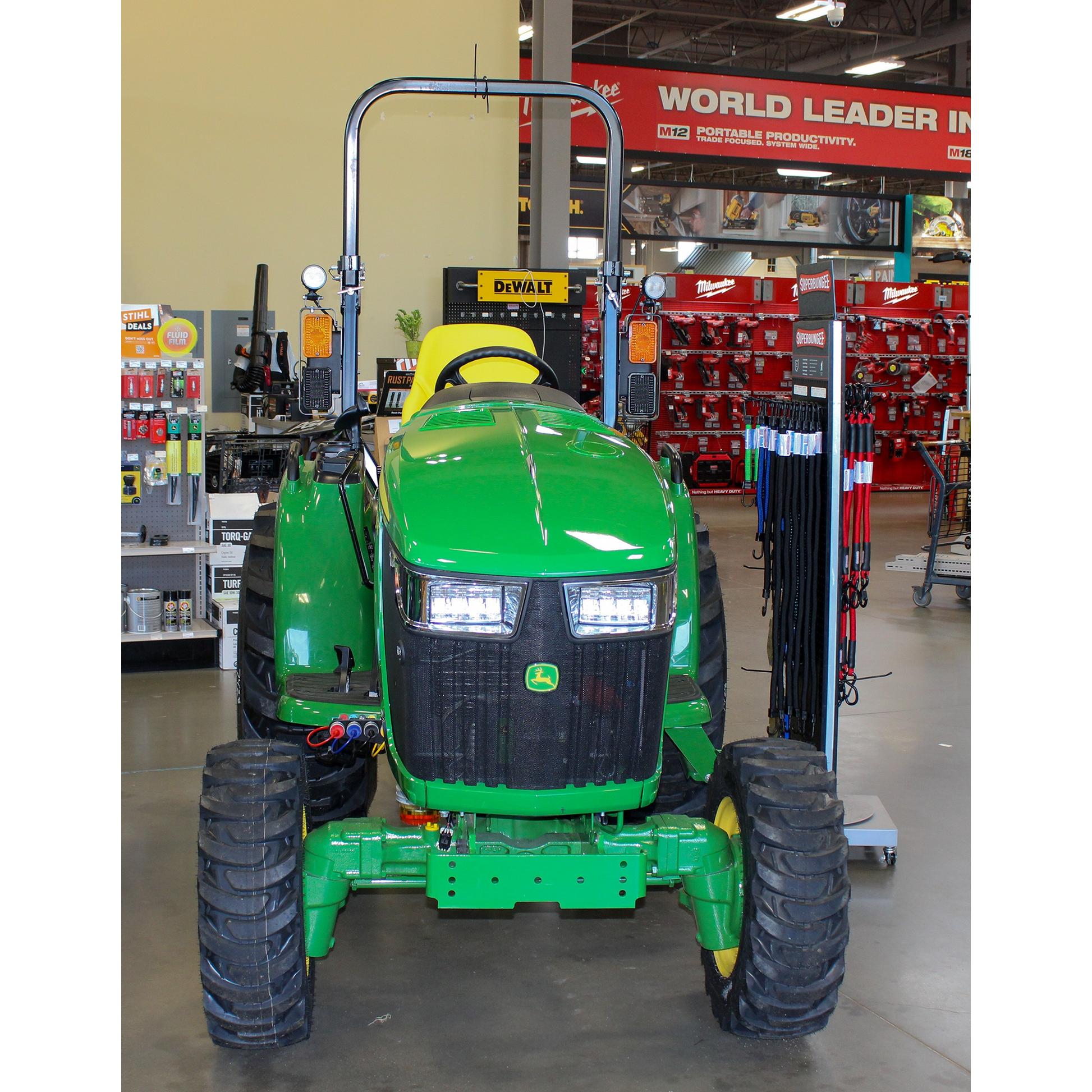 Front view of a green John Deere compact utility tractor with yellow seat and large tread tires displayed indoors among outdoor power equipment and tools in a hardware store.