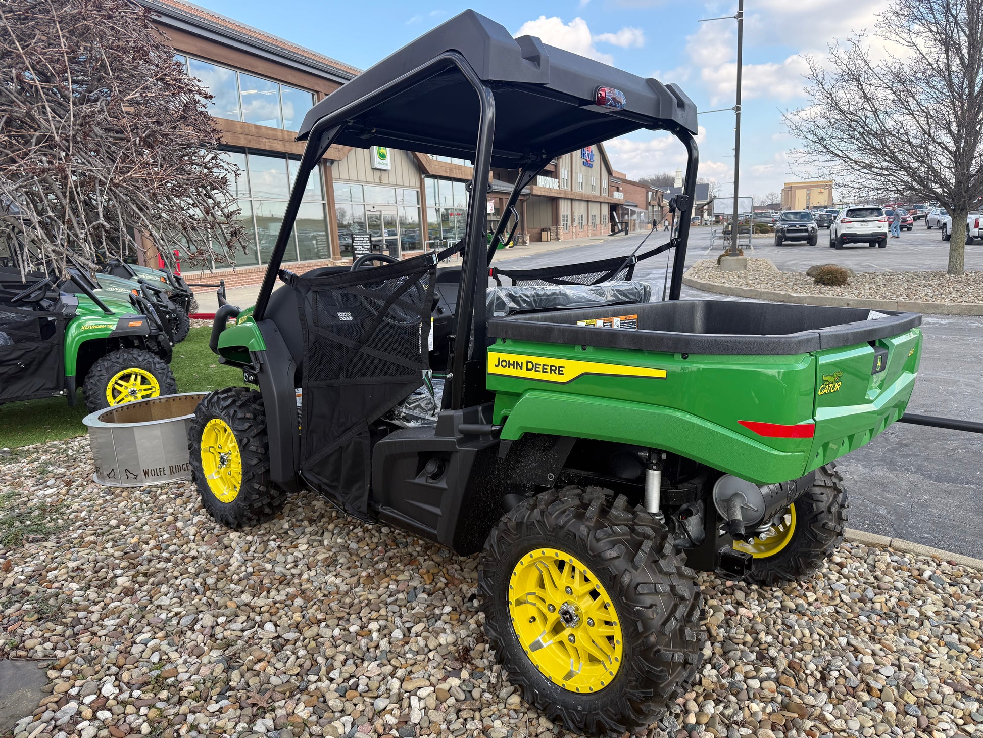 Green John Deere mid-size crossover utility vehicle with yellow wheels and cargo bed, parked on gravel outside a Hartville Hardware store.