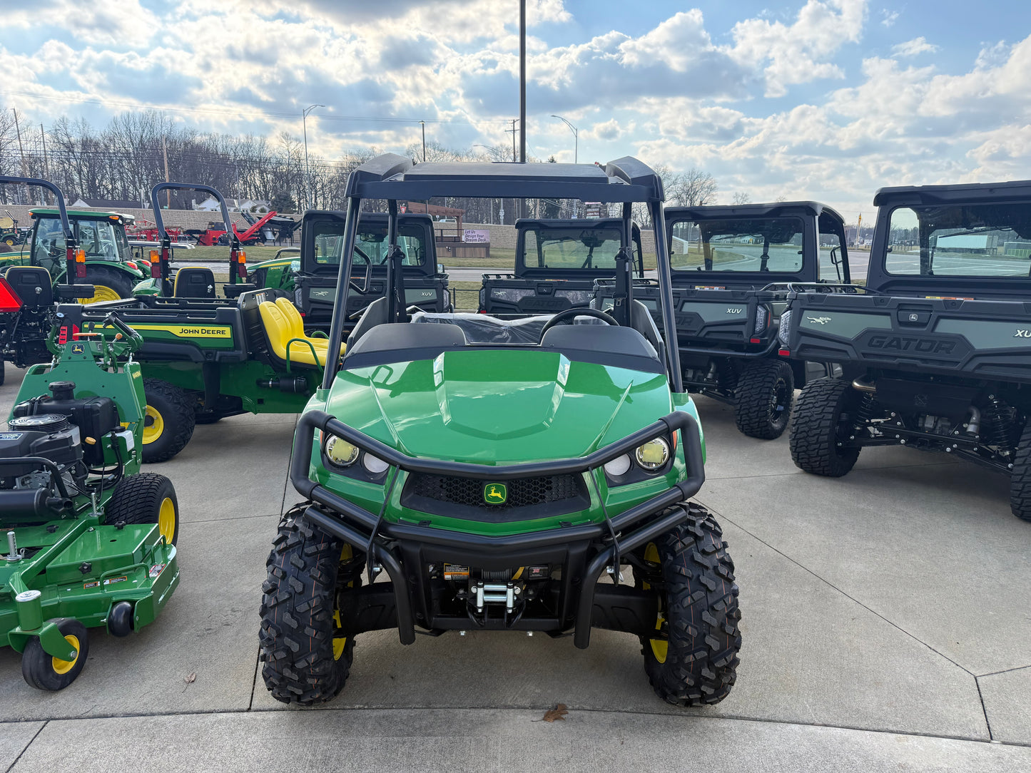 Green mid-size John Deere utility vehicle with rugged off-road tires parked outdoors among other utility vehicles and equipment in a dealership lot.