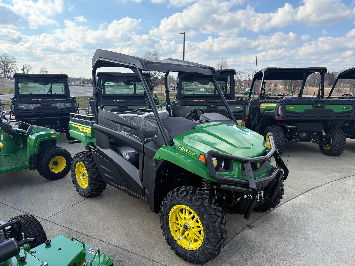 Green and yellow mid-size John Deere crossover utility vehicle with off-road tires displayed outdoors among other John Deere UTVs and equipment.