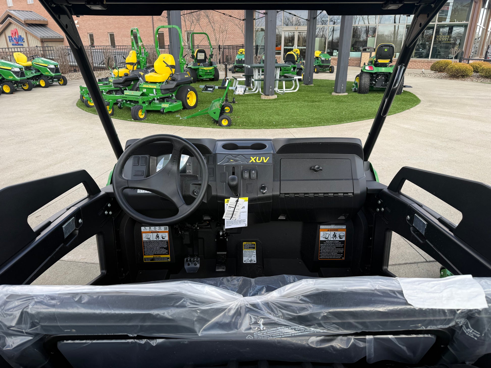 Interior view of a John Deere mid-size crossover utility vehicle featuring a steering wheel, dashboard controls, and a protective plastic-covered bench seat in a dealership lot.