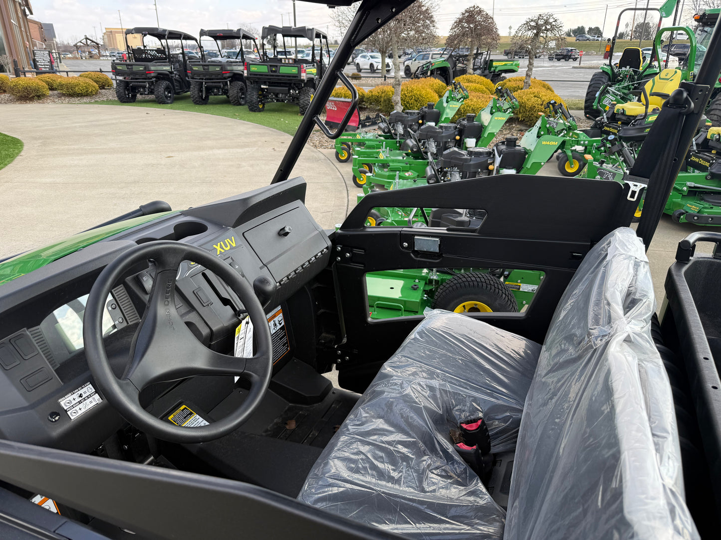 Interior view of a John Deere mid-size crossover utility vehicle showing the steering wheel, dashboard, and a bench seat covered in protective plastic at a dealership lot.