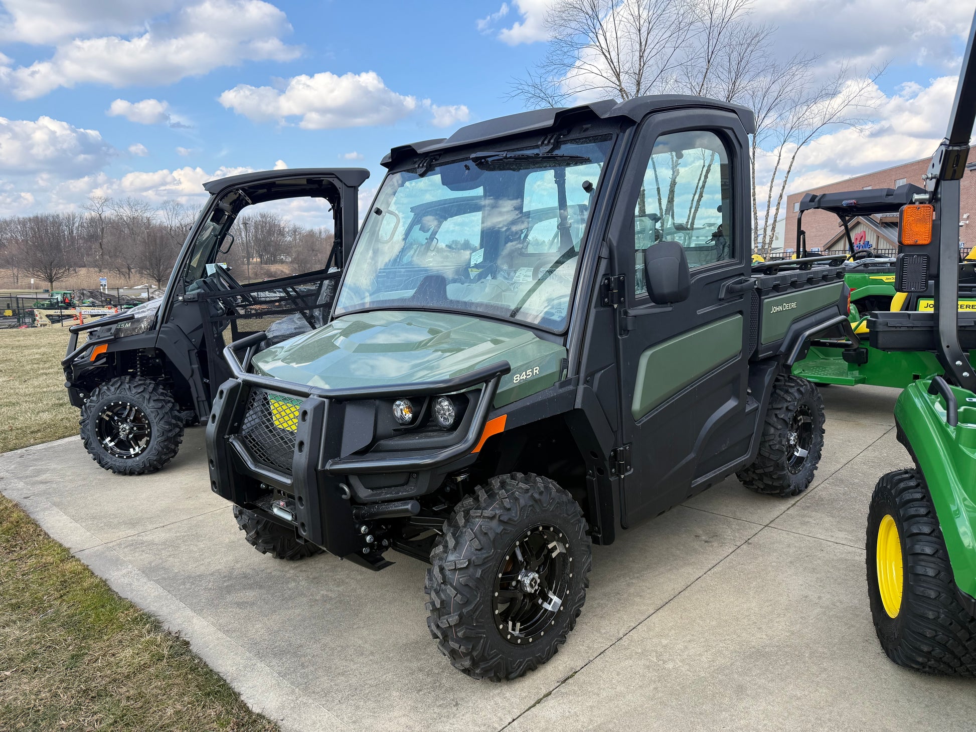 Green John Deere crossover utility vehicle with enclosed cab, rugged off-road tires, and cargo bed parked on concrete near other equipment at dealership lot