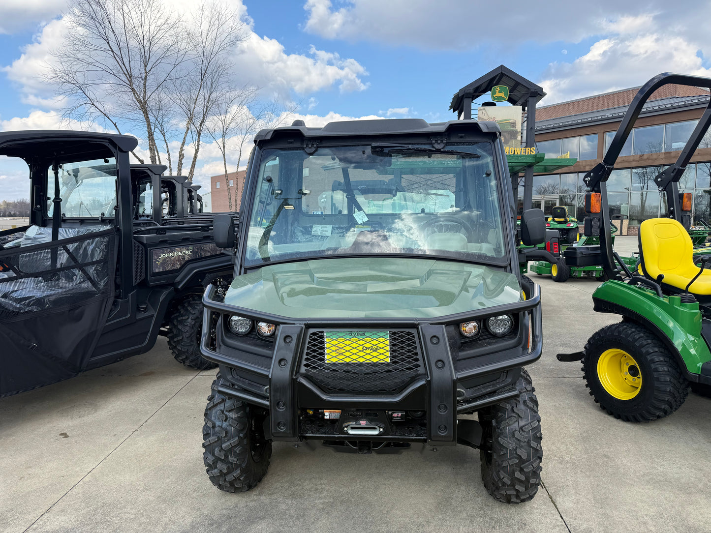 Front view of a green John Deere utility vehicle with off-road tires, protective grille, and enclosed cab parked on a concrete lot among other equipment