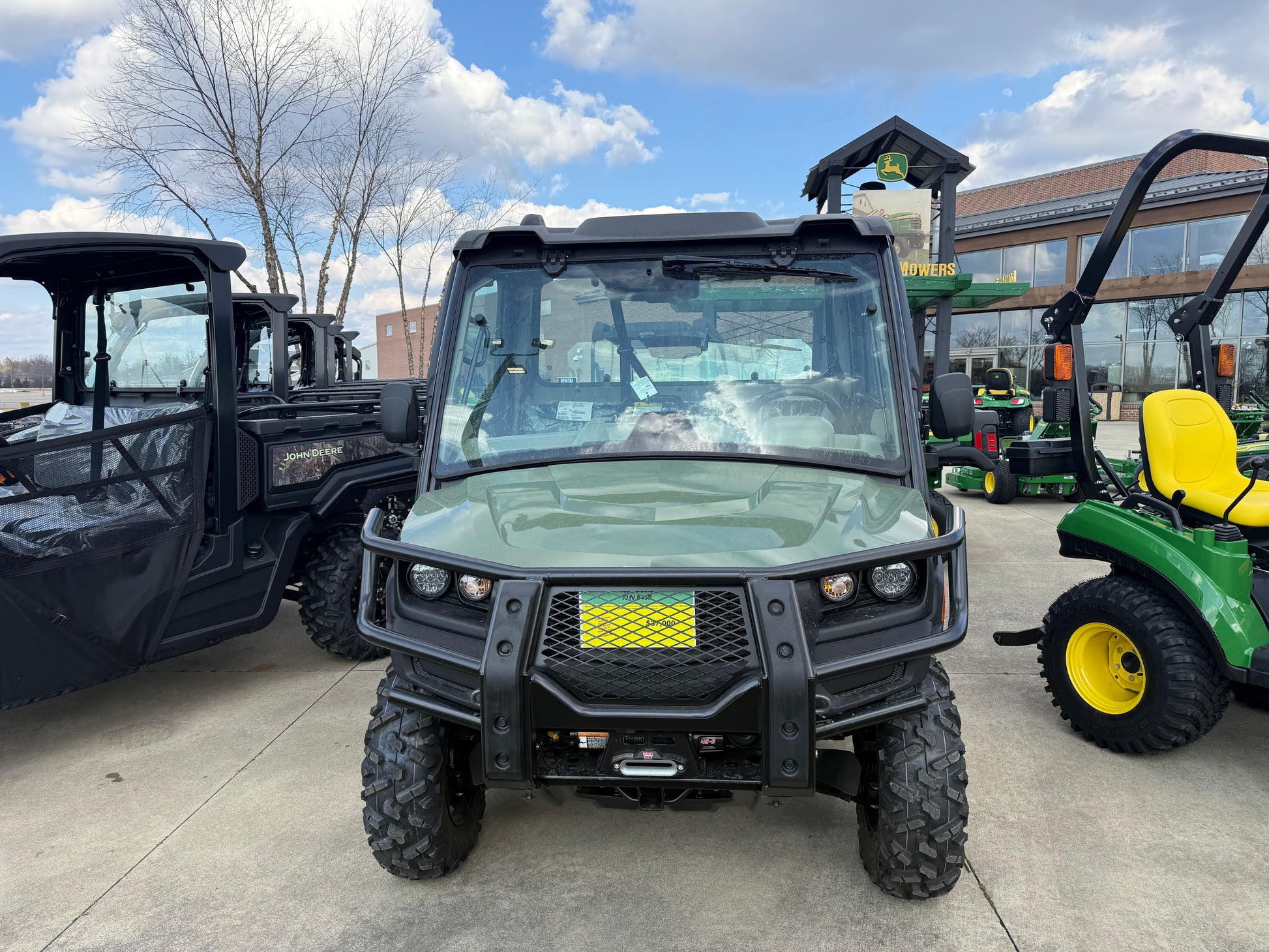 Front view of a green John Deere utility vehicle with off-road tires, protective grille, and enclosed cab parked on a concrete lot among other equipment