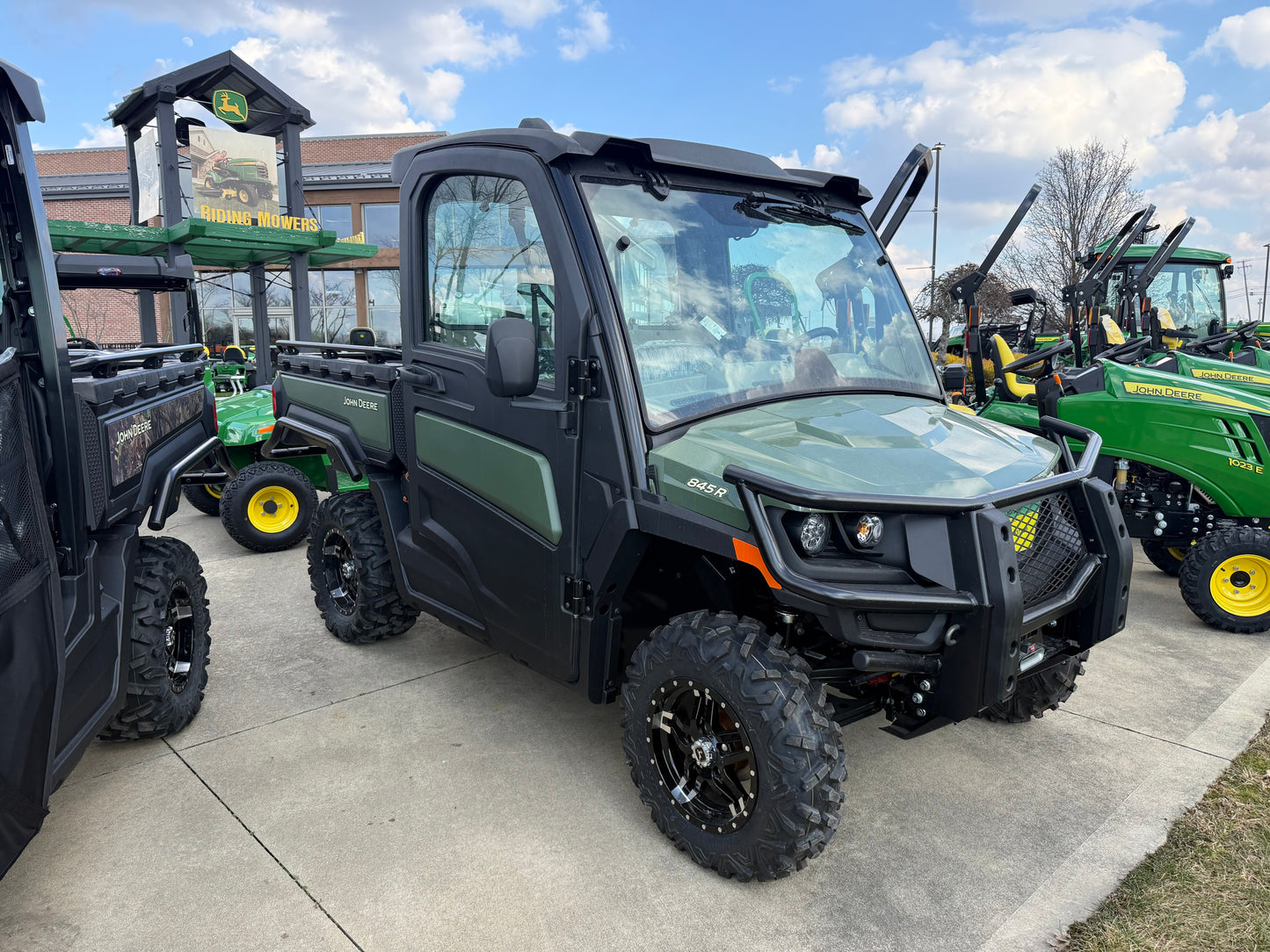 Green John Deere Custom XUV 845R crossover utility vehicle with enclosed cab, rugged tires, and cargo bed displayed outdoors at dealership among other equipment