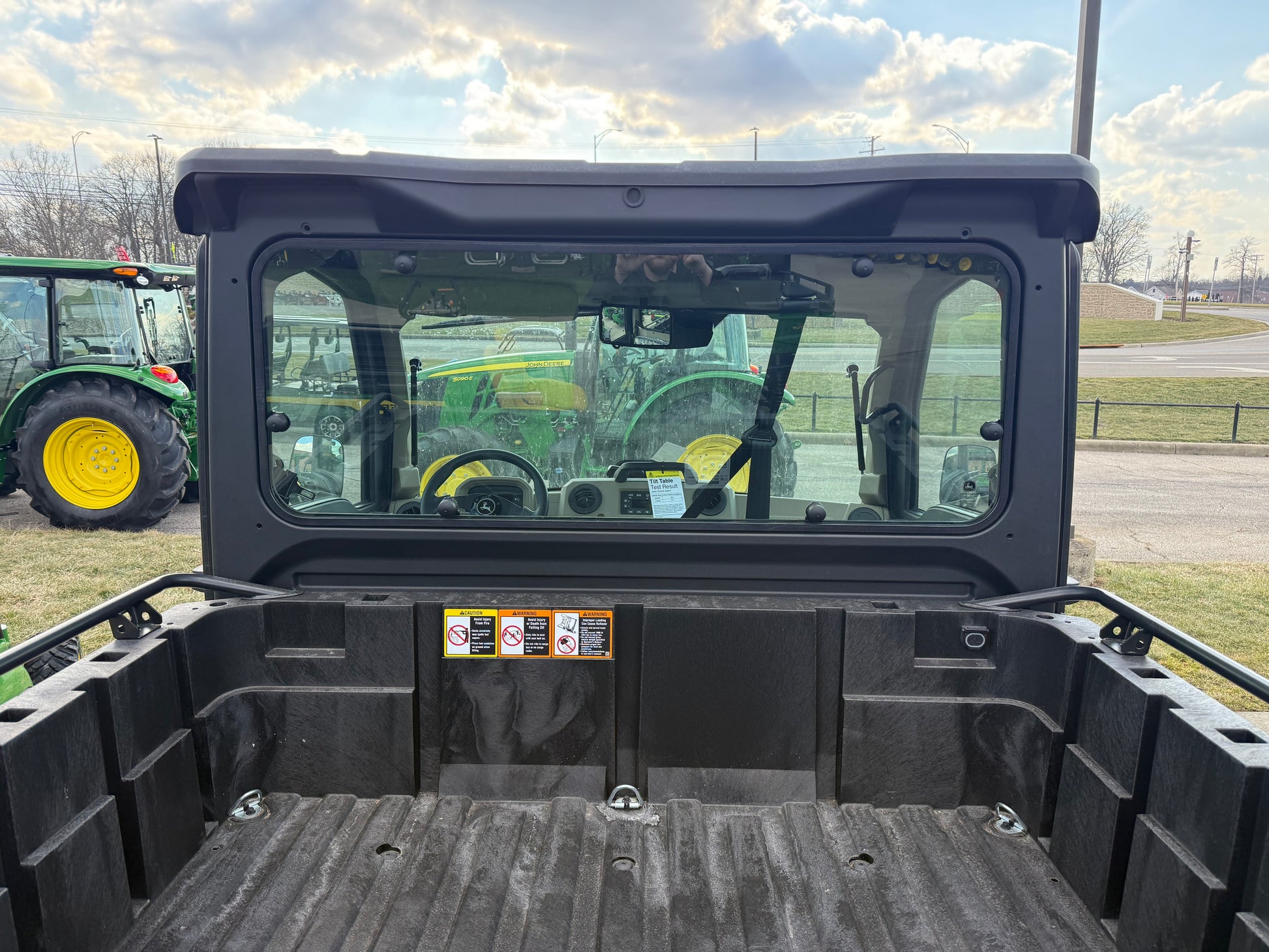 Rear cargo bed and enclosed cab view of a John Deere Custom XUV 845R crossover utility vehicle parked outdoors near other John Deere equipment at dealership lot