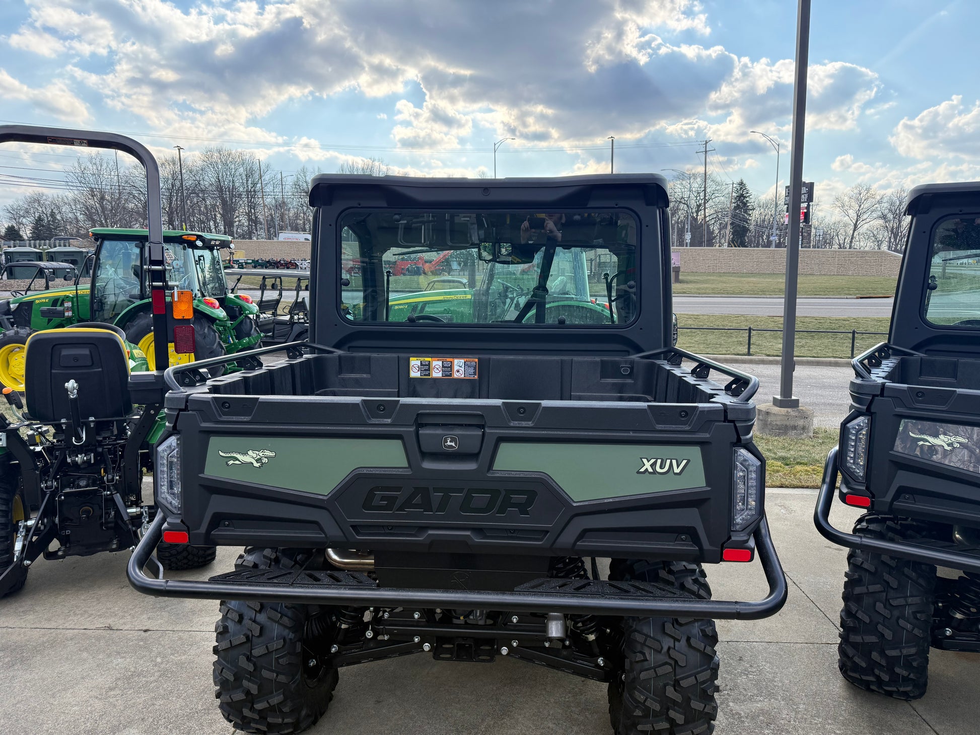 Rear view of a rugged John Deere green crossover utility vehicle with cargo bed and protective cab, parked outdoors near other equipment at dealership lot