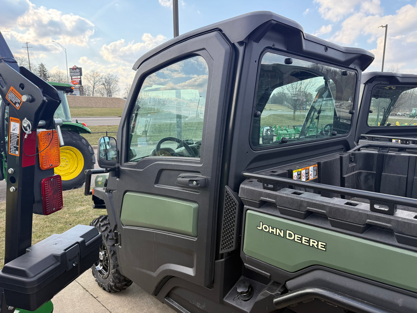 Close-up side view of a John Deere crossover utility vehicle with enclosed cab, showing rugged cargo bed and part of the interior at outdoor equipment lot