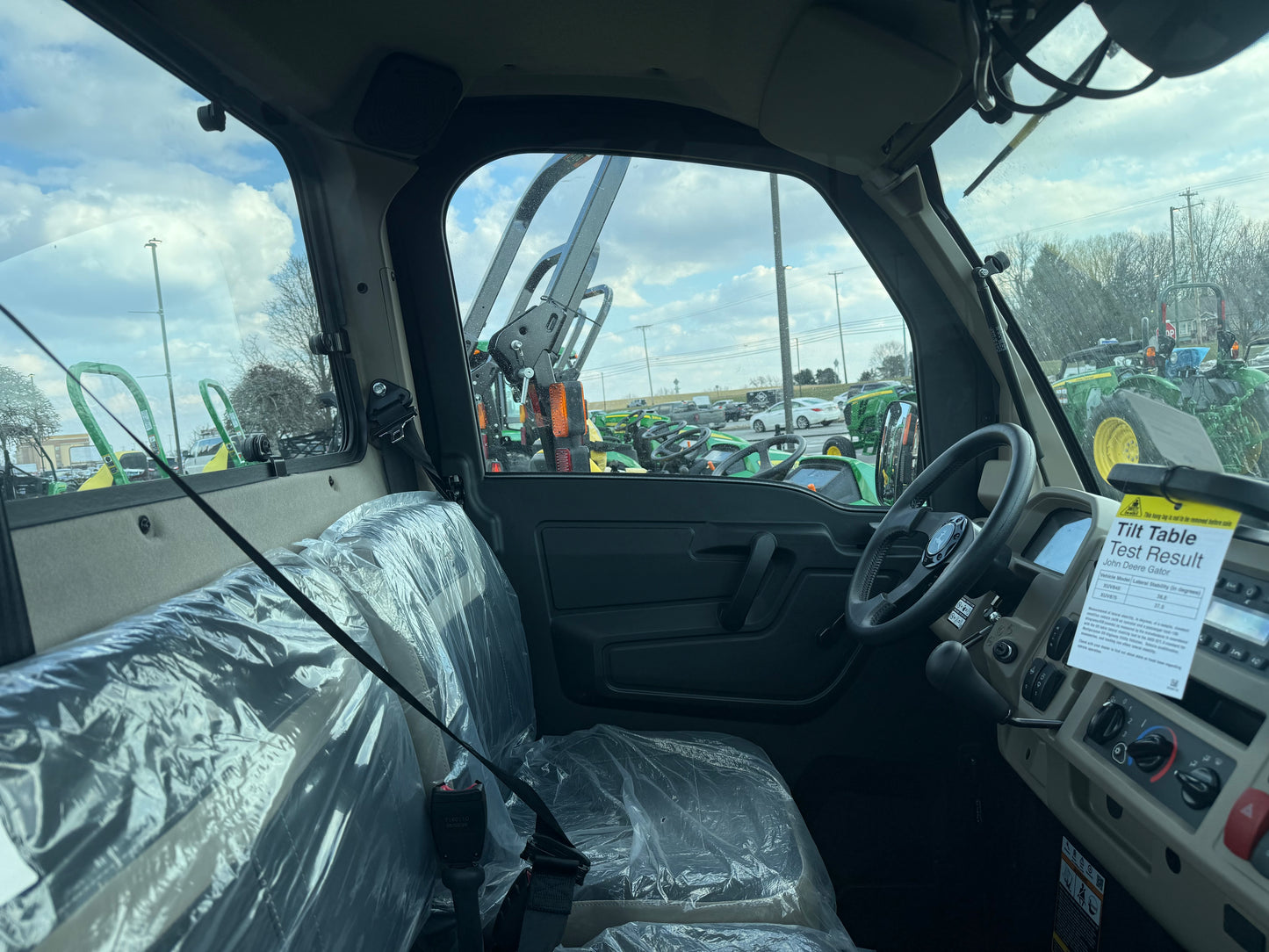 Interior view of a John Deere utility vehicle showing steering wheel, dashboard controls, and protective plastic-covered bench seat at dealership lot