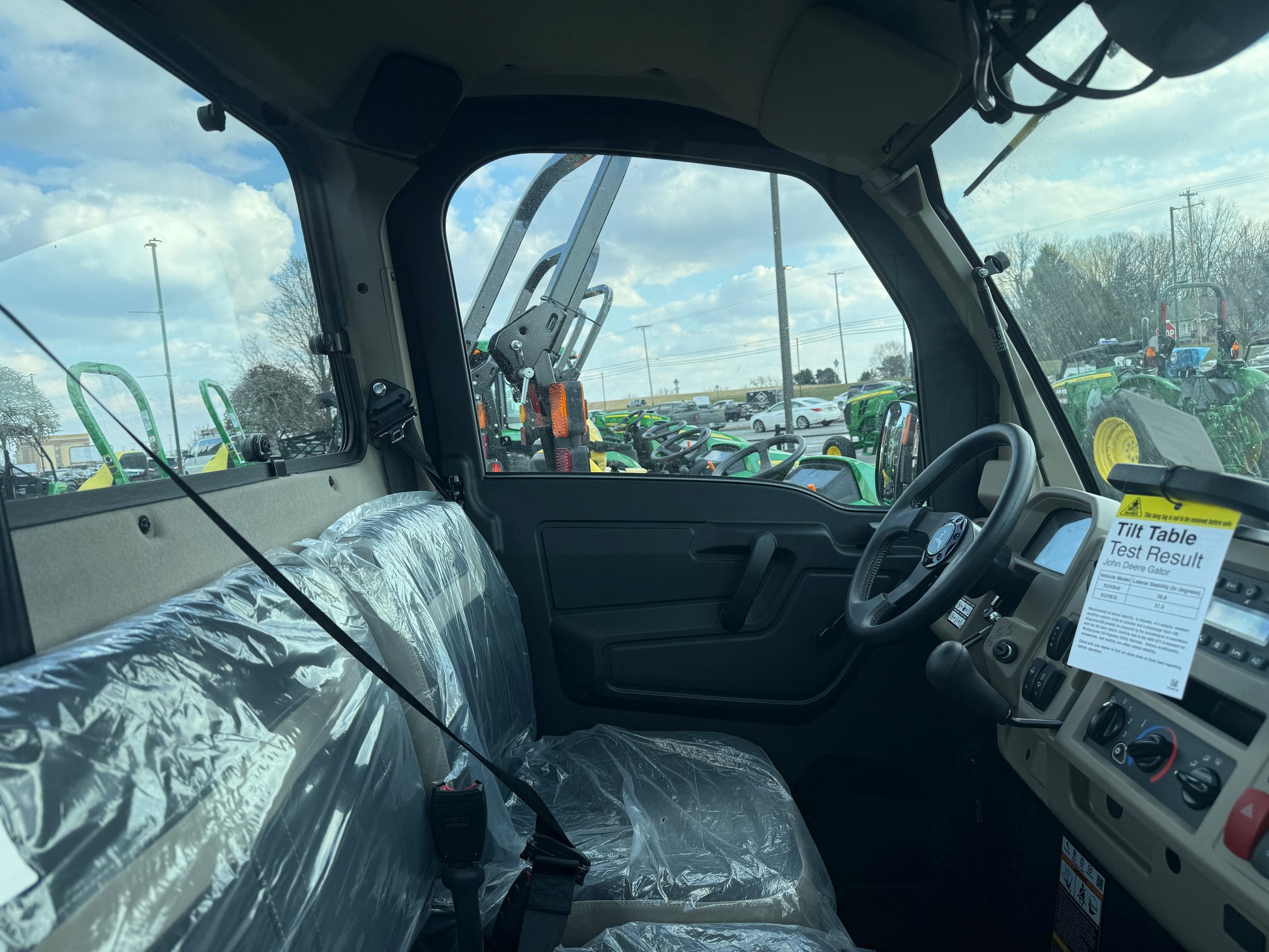 Interior view of a John Deere utility vehicle showing steering wheel, dashboard controls, and protective plastic-covered bench seat at dealership lot