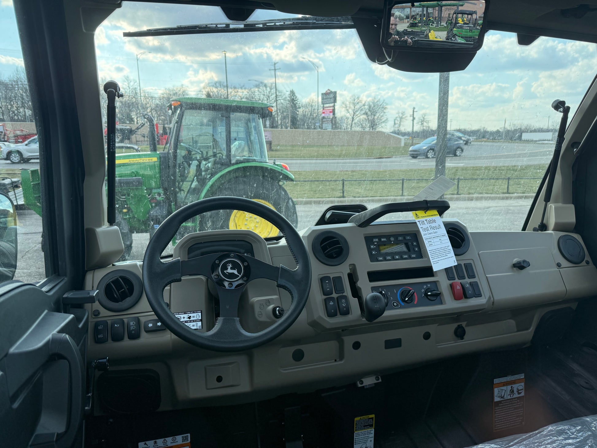 View of the dashboard and steering wheel inside a John Deere JD697731 Custom XUV 845R utility vehicle at an outdoor equipment lot with a tractor visible through the windshield.