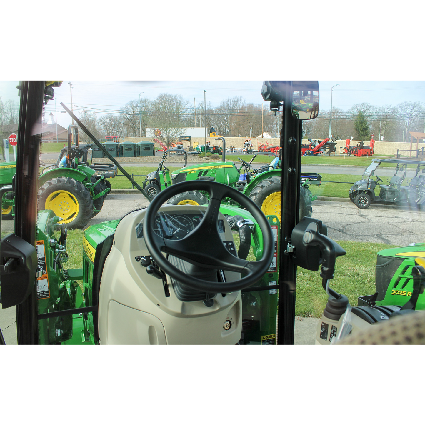View from inside a tractor cab showing steering wheel and controls, with several green John Deere tractors and outdoor power equipment visible outside on display.