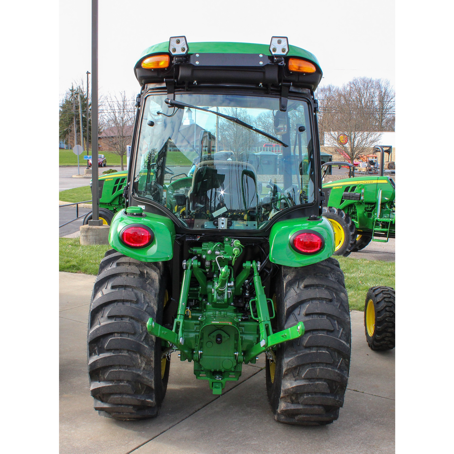 Rear view of a green John Deere compact tractor with rugged tires and enclosed cab, displayed outdoors among other outdoor power equipment and tools.