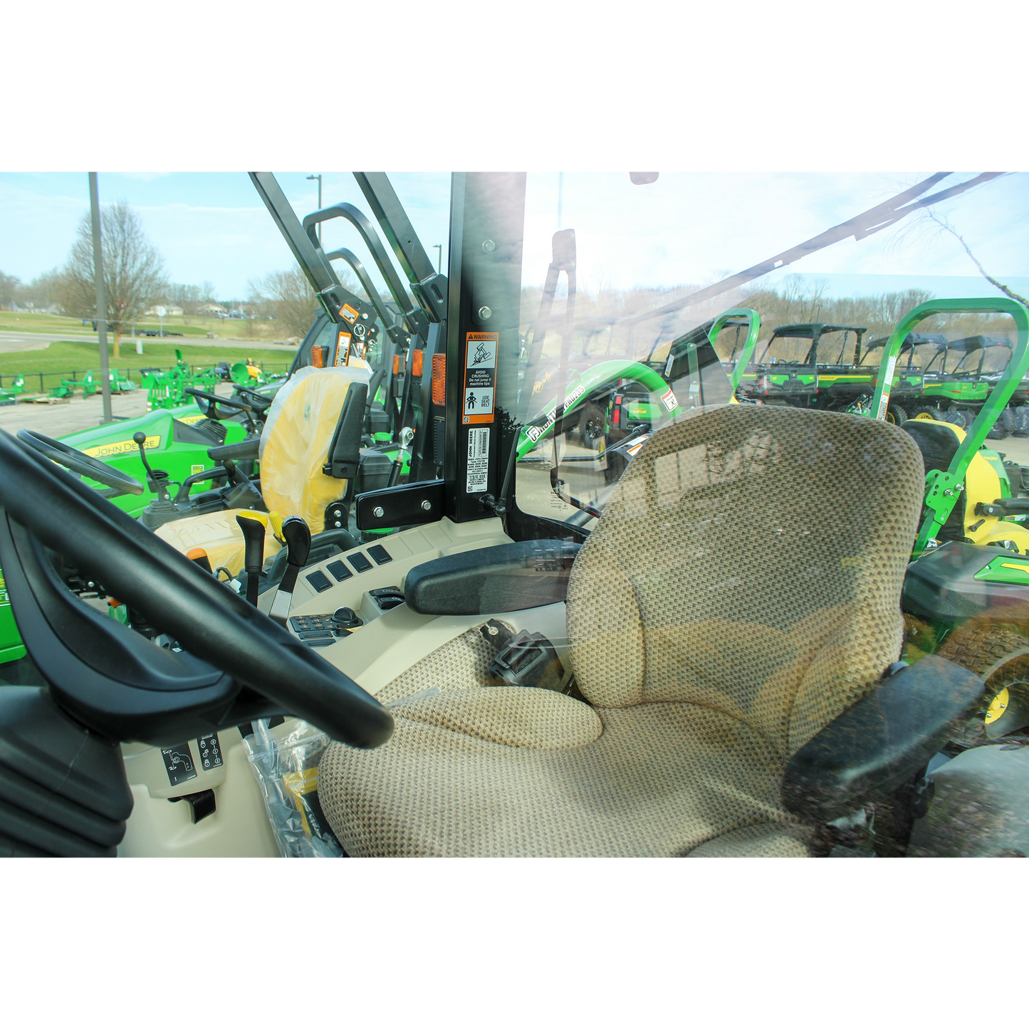Interior view of a beige operator seat and control panel inside a John Deere compact tractor cab, with outdoor power equipment visible through the windows.