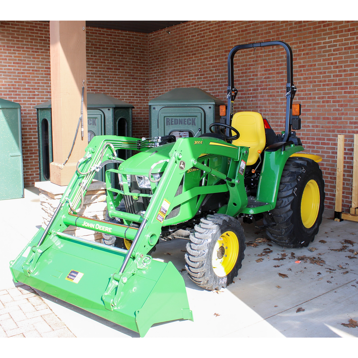 Green John Deere compact utility tractor with front loader and yellow seat, displayed indoors among outdoor power equipment and shop tools.