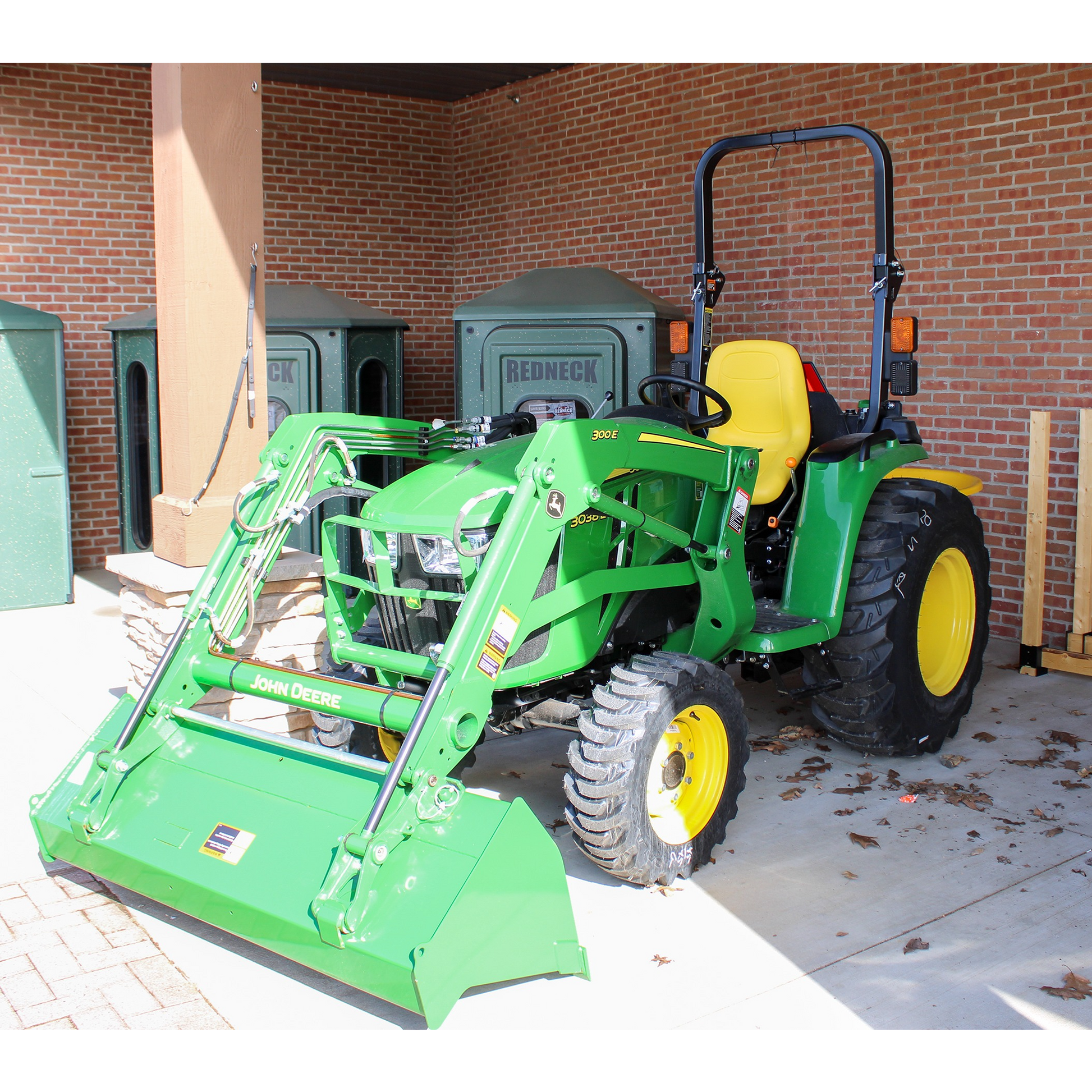 Green John Deere compact utility tractor with front loader and yellow seat, displayed indoors among outdoor power equipment and shop tools.