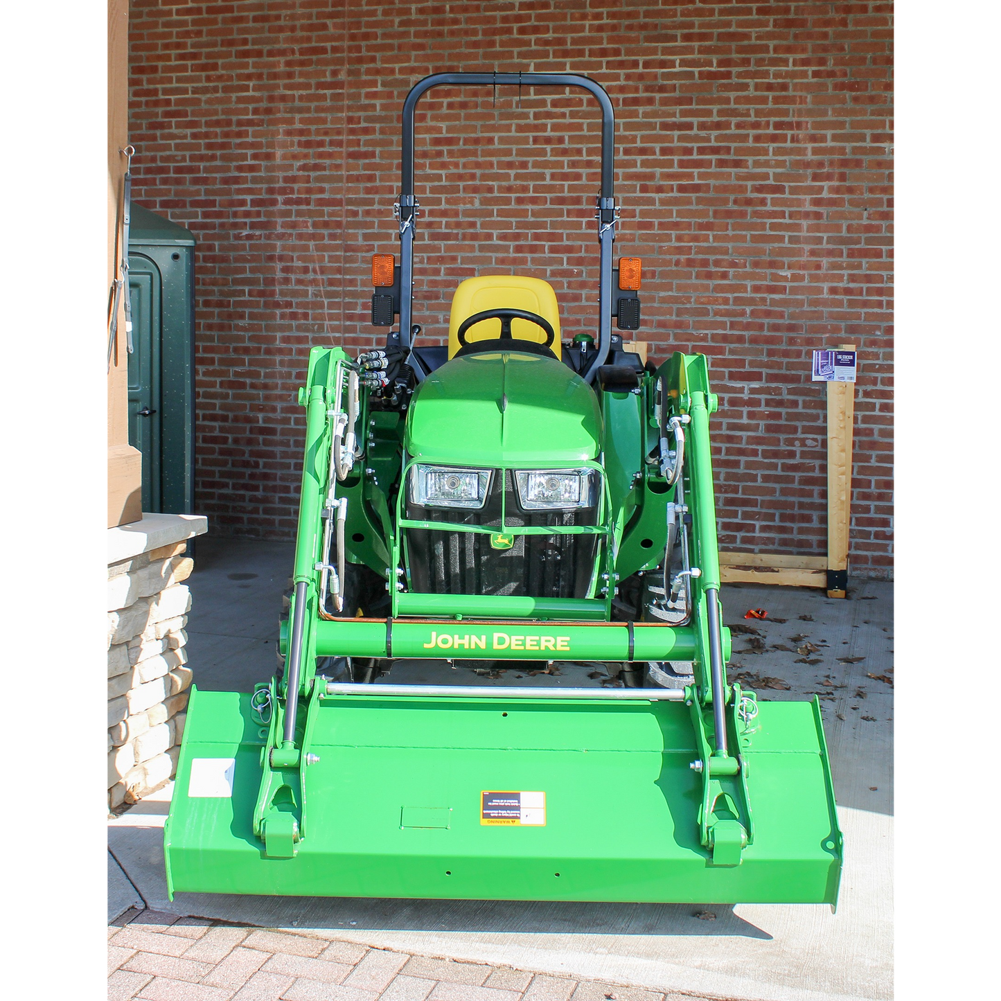 Front view of a green John Deere compact utility tractor with loader attachment and yellow operator seat, displayed indoors with brick wall backdrop for Outdoor Power Equipment collection.