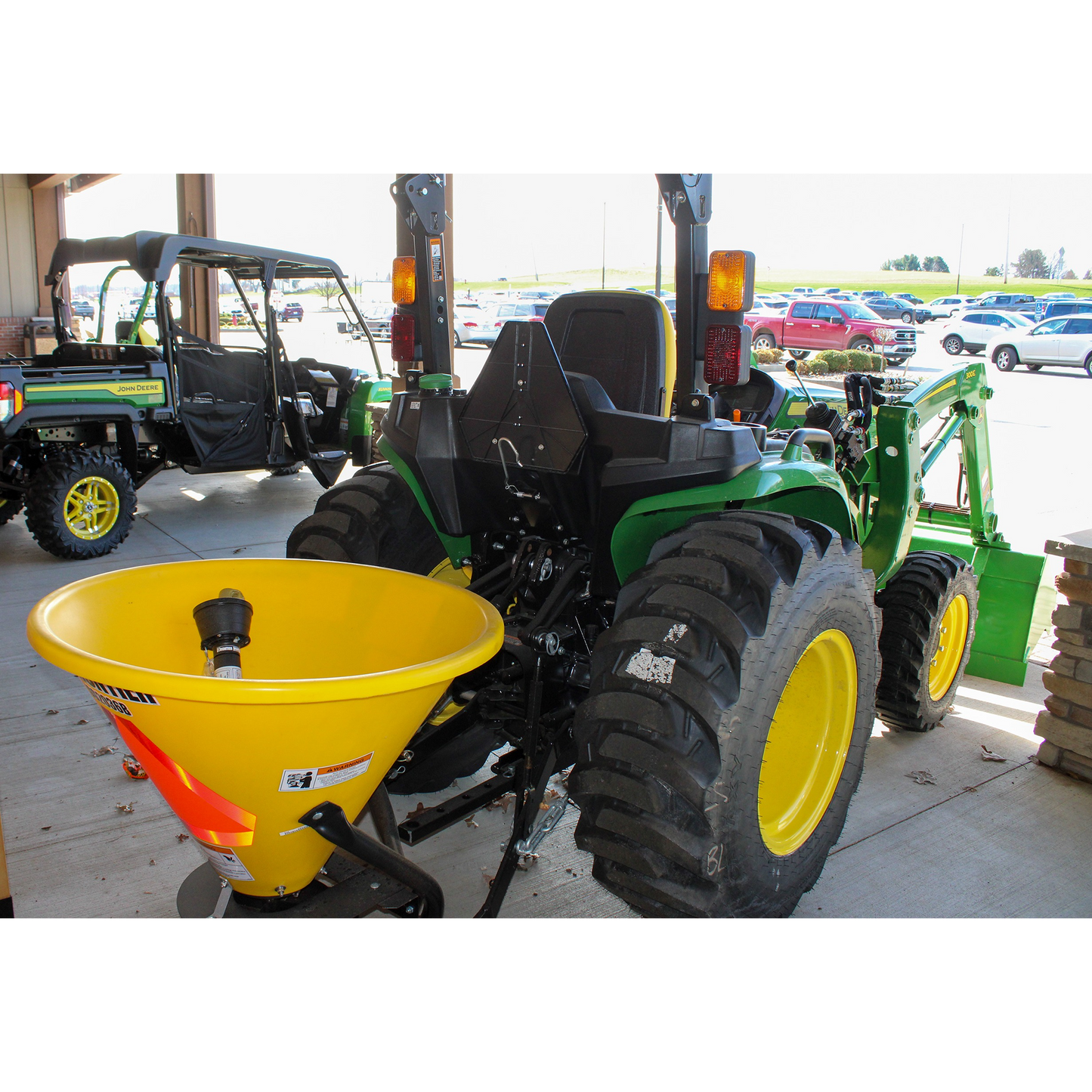 Rear view of a green John Deere compact utility tractor equipped with a yellow fertilizer spreader attachment, displayed among outdoor power equipment at a Hartville Hardware store.