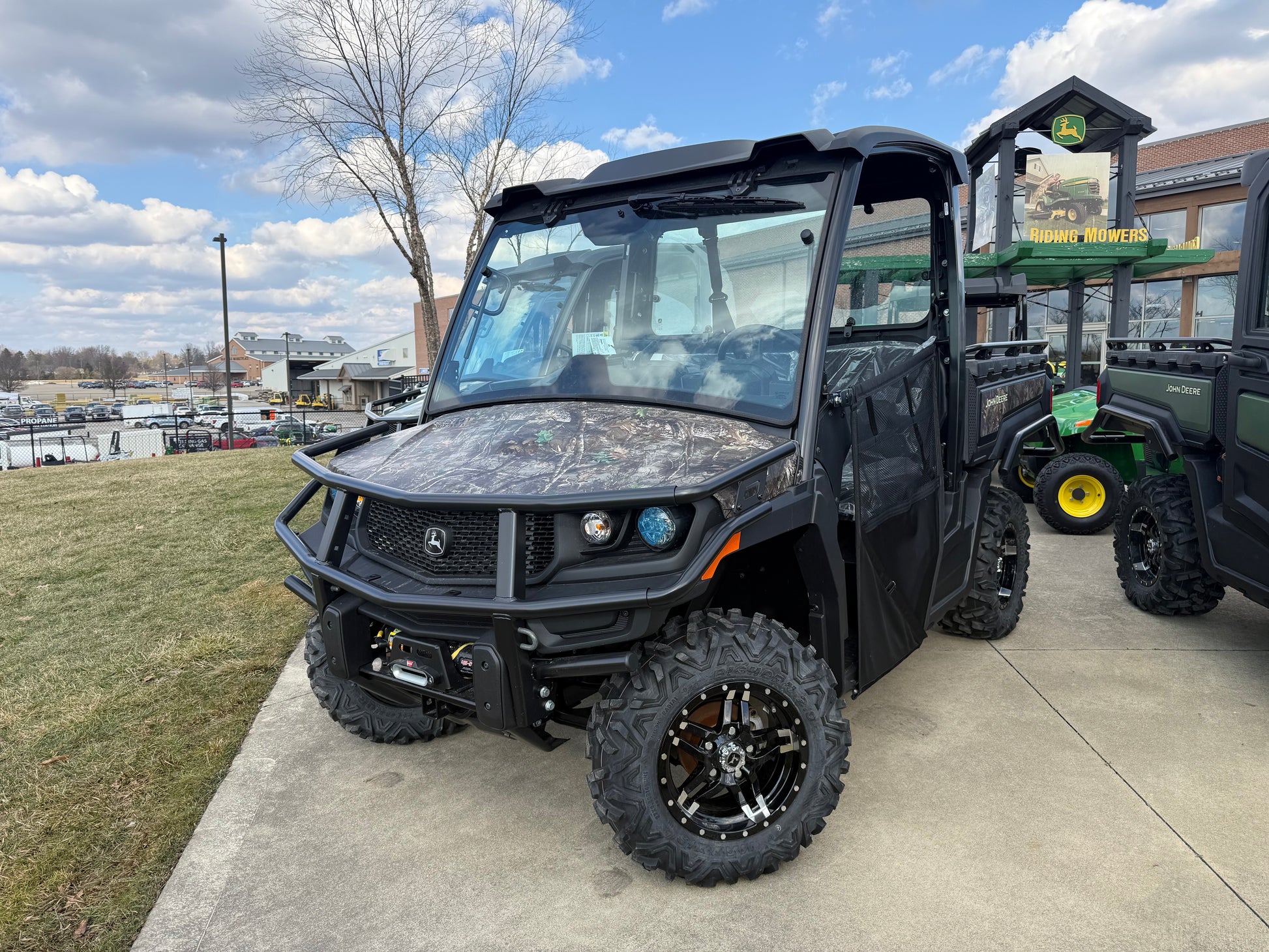 John Deere Custom XUV 845M crossover utility vehicle with camouflage hood, enclosed cab, and rugged off-road tires displayed outdoors at dealership lot among John Deere equipment.