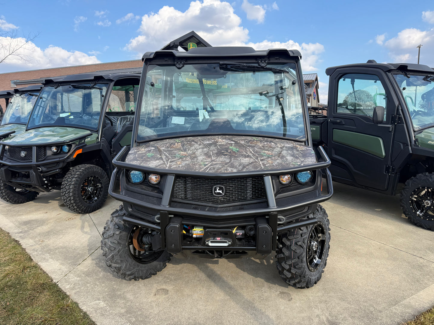 Front view of a John Deere utility vehicle with camo hood, protective grille, and rugged tires parked outdoors among other John Deere UTVs and equipment.
