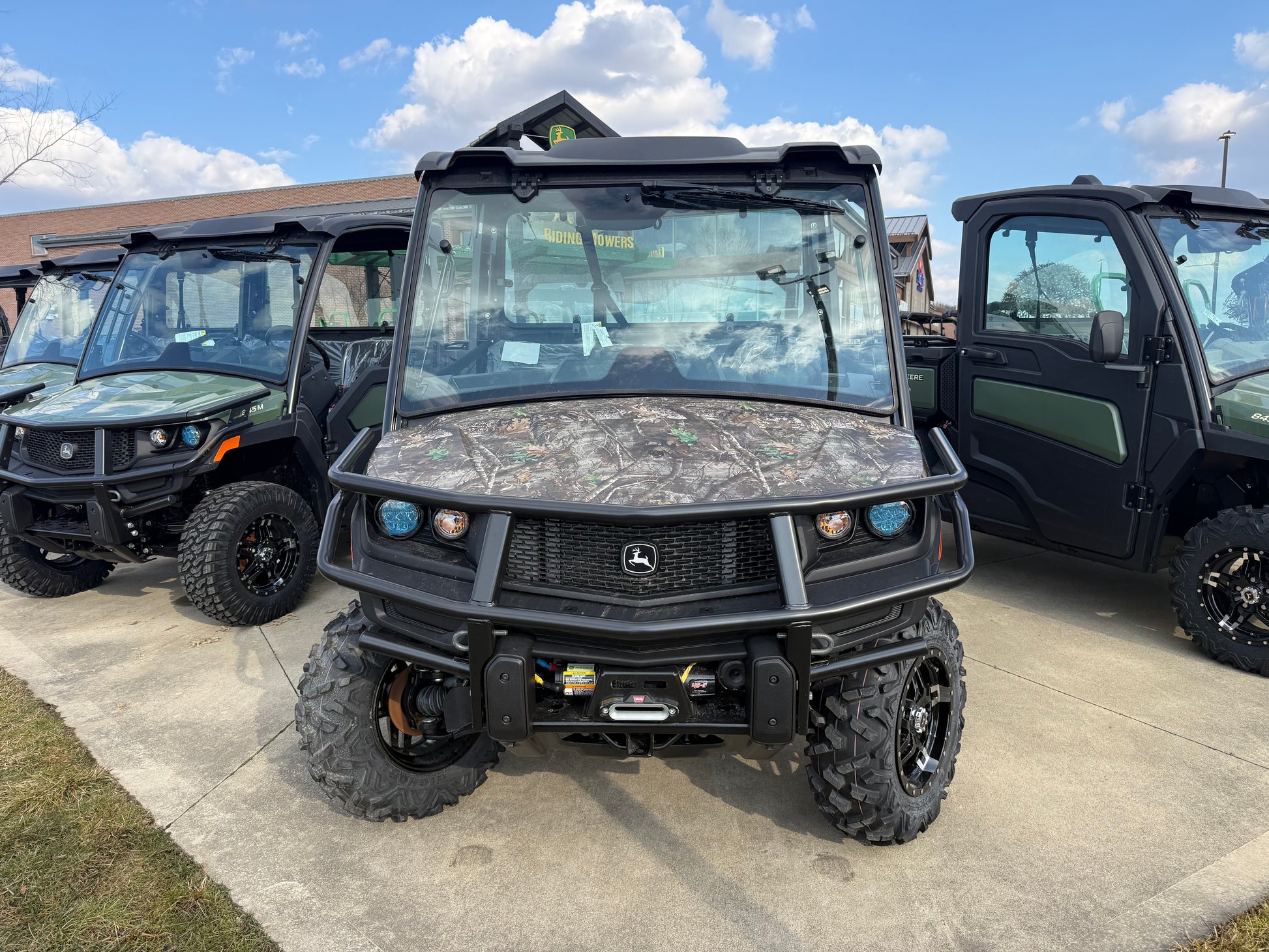 Front view of a John Deere utility vehicle with camo hood, protective grille, and rugged tires parked outdoors among other John Deere UTVs and equipment.