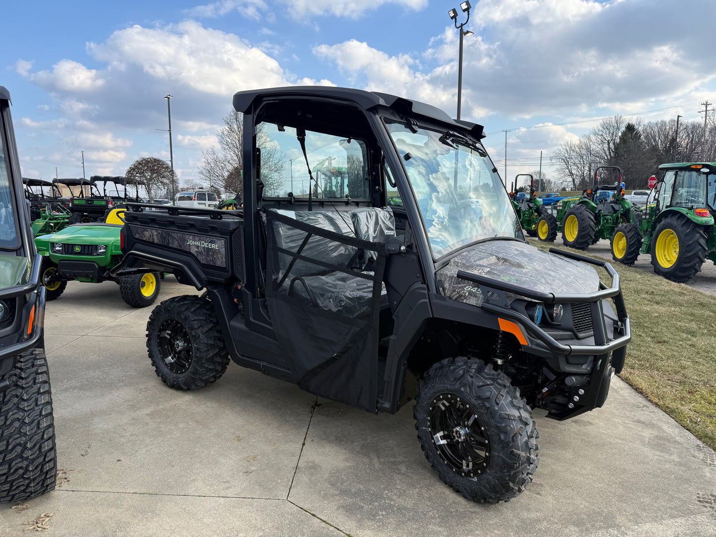 Side view of a black John Deere Custom XUV 845M crossover utility vehicle with enclosed cab and rugged off-road tires parked on concrete at an outdoor dealership lot among other green John Deere equipment