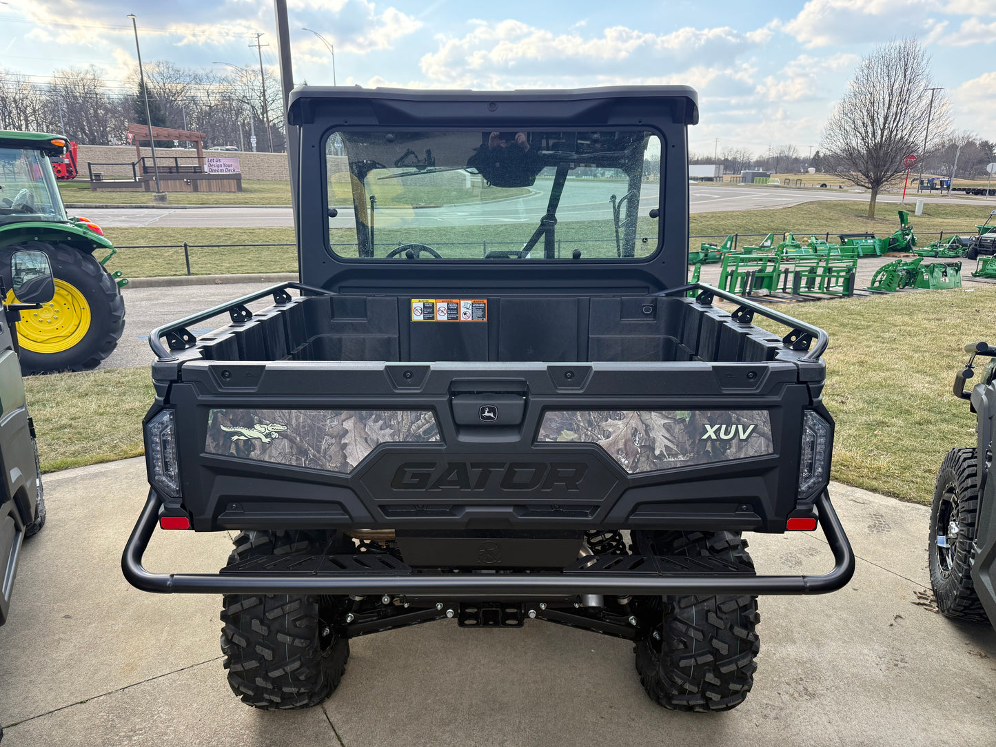Rear view of a John Deere crossover utility vehicle featuring a spacious cargo bed with camo accents and protective frame at an outdoor dealership lot