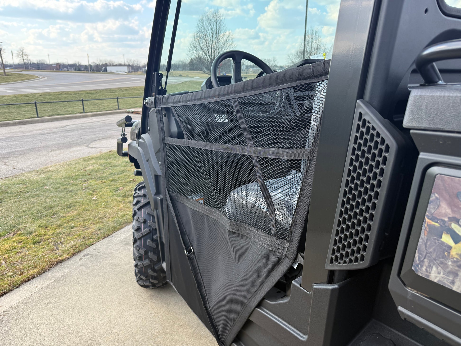 Close-up side view of a John Deere JD718267 Custom XUV 845M crossover utility vehicle showing mesh door panel and rugged tire at outdoor location near a road and grassy area.